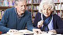 Eine alte Frau und ein alter Mann sitzen an einem Tisch und lesen zusammen ein Buch.