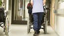 Eine Pflegekraft, von hinten zu sehen, schiebt einen älterne Menschen im Rollstuhl über einen Flur .