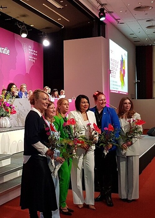 Eine Gruppe Frauen mit Blumensträußen in der Hand steht in einer Kongresshalle.