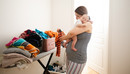 Young mother holding sleepeing baby on her shoulder and trying to sort the laundry simultaniously. Mother combines everyday routines with raising her newborn baby.