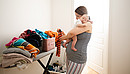 Young mother holding sleepeing baby on her shoulder and trying to sort the laundry simultaniously. Mother combines everyday routines with raising her newborn baby.