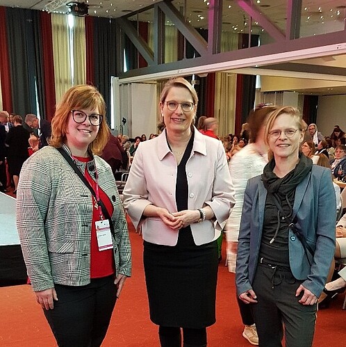 Drei Frauen stehen in einer Kongresshalle und blicken in die Kamera