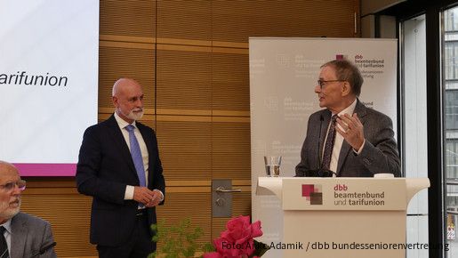 Zwei Männer in Anzügen unterhalten sich auf einer Bühne.