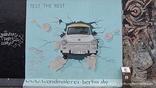 Malerei auf der Berliner Mauer, wie ein altes Auto die Mauer durchbricht