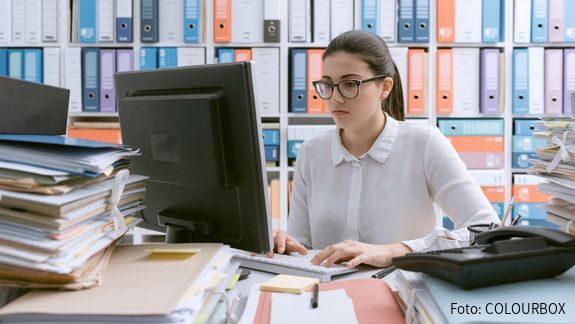 Verwaltung Junge Behördenmitarbeiterin sitzt am Schreibtisch hinter einem Berg von Akten, auch im Hintergrund stapeln sich Akten im Regal.