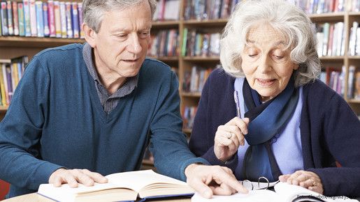 Eine alte Frau und ein alter Mann sitzen an einem Tisch und lesen zusammen ein Buch.
