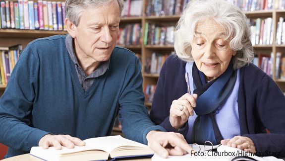 Eine alte Frau und ein alter Mann sitzen an einem Tisch und lesen zusammen ein Buch.