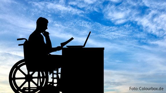 Die Silhouette eines Mannes im Rollstuhl, der an einem Schreibtisch sitzt und arbeitet.