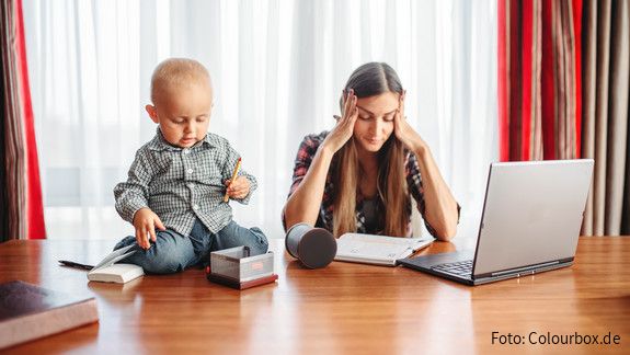 Erwerbsarbeit und private Care-Arbeit müssen zusammen gedacht werden Eine sichtlich gestresste Mutter versucht zu Hause am Laptop zu arbeiten, während neben ihr auf dem Schreibtisch ein Kleinkind sitzt und mit Büroutensilien spielt.