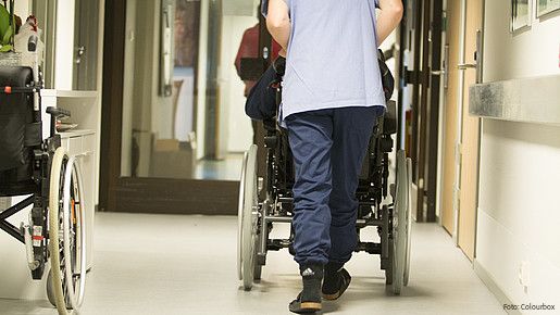 Eine Pflegekraft, von hinten zu sehen, schiebt einen älterne Menschen im Rollstuhl über einen Flur .