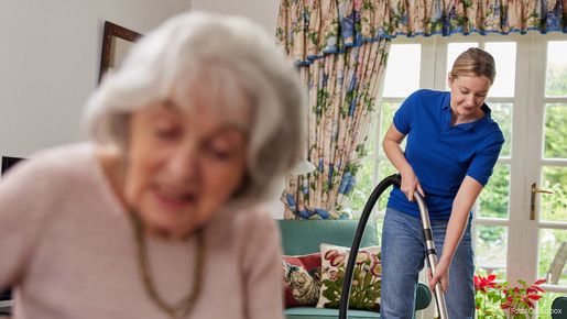 Im Vordergrund sitzt eine Senioren, im Hintergrund ist ihre Haushaltshilfe zu sehen, die gerade stabsaugt.