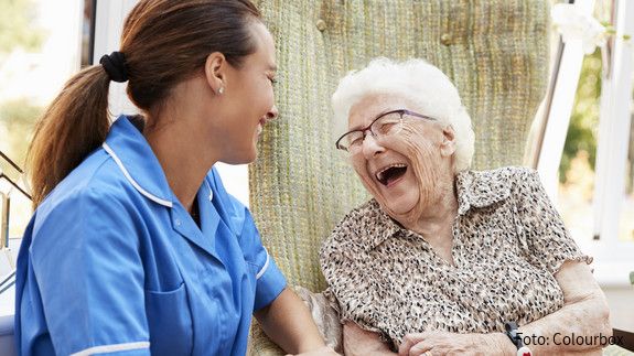 1. Oktober: Internationaler Tag der älteren Menschen Szene aus Seniorenresidenz: Eine Seniorin sitzt auf einer Bank, neben ihr eine Pflegefachkraft. Beide lachen.