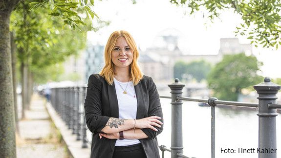 Karoline Herrmann Eine junge Frau steht mit verschränkten Armen draußen und lächelt in die Kamera.