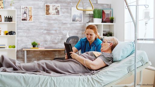 Eine ältere Frau liegt im Bett und eine Pflegerin hilft ihr dabei, ein Tablet zu bedienen