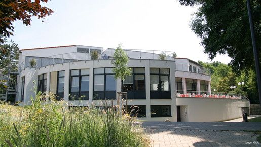 Ein großes, helles, rundliches Haus ist im Mittelpunkt des Bildes. Im linken Vordergrund ist eine hochwachsende Wiese zu sehen.