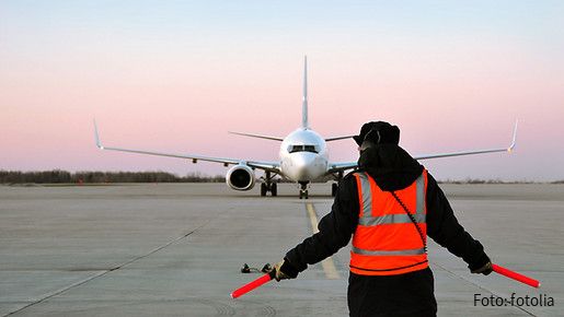Bodenverkehrsdienste Ein Fluglotse in einer roten Warnweste steht mit dem Rücken zur Kamera. Weit vor ihm ist ein Flugzeug zu sehen, das auf der Flugbahn steht.